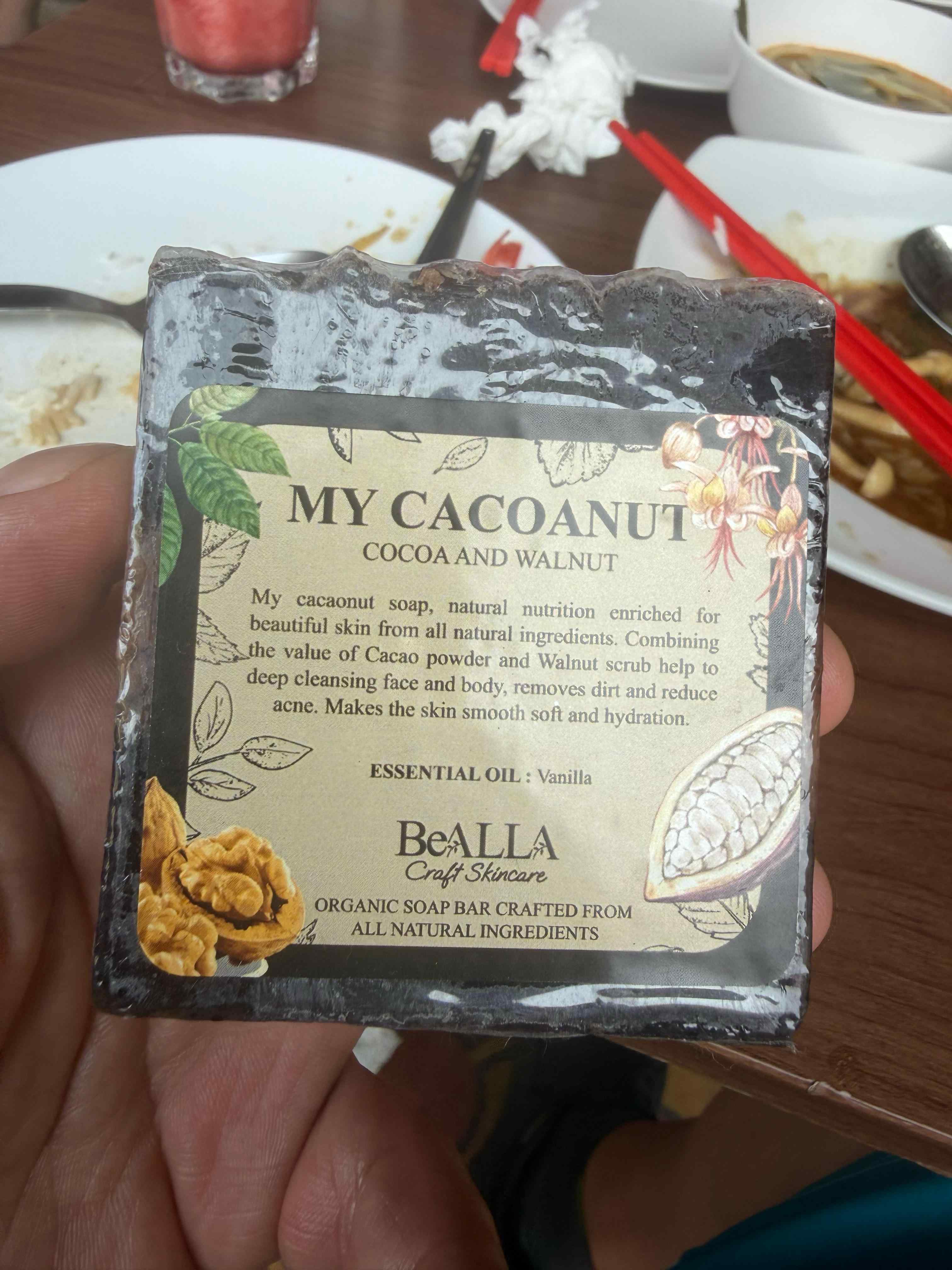 Bar of soap labeled 'My CacaoNut' held in front of a blurred background with food items.